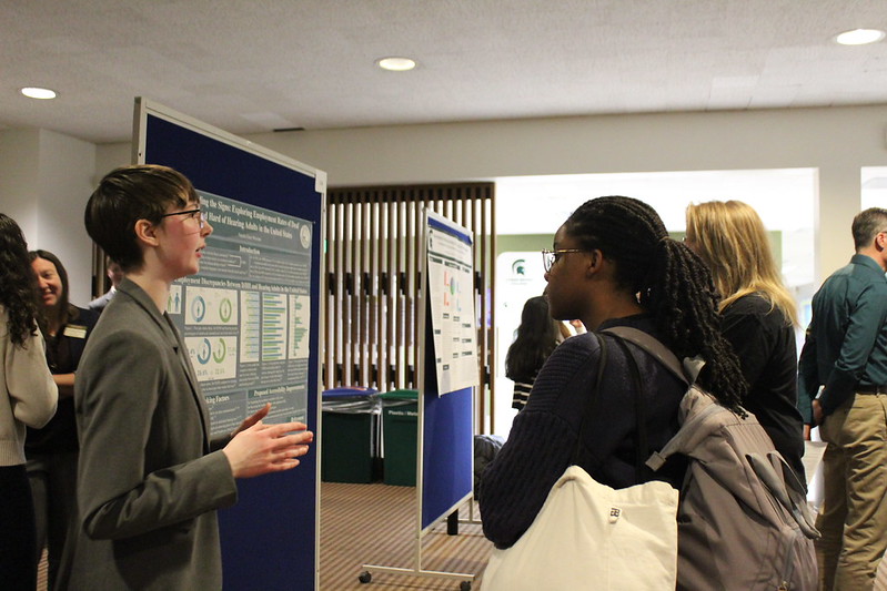 A presenter discusses their research poster with an audience member. In the background there are four or five other presenters and audience members
